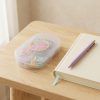 Estuche de clips y pinzas pastel sobre una mesa de madera junto a un cuaderno y un bolígrafo