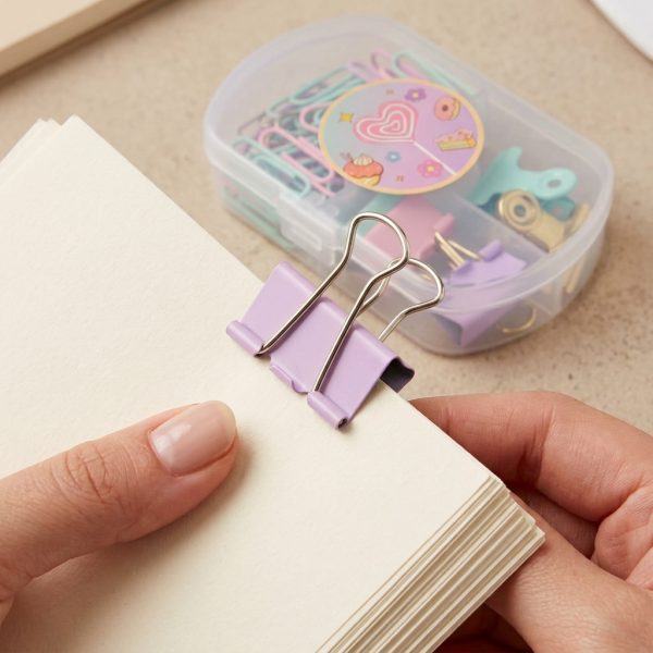 Manos sosteniendo hojas sujetas con una pinza lila de asas doradas, con el estuche al fondo