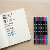 Set de rotuladores YUWEI Amazcolor junto a un cuaderno con ejercicios de caligrafía sobre una mesa de madera.