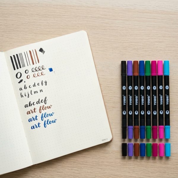 Set de rotuladores YUWEI Amazcolor junto a un cuaderno con ejercicios de caligrafía sobre una mesa de madera.