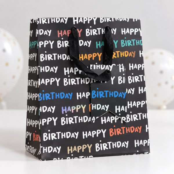Bolsa negra Happy Birthday sobre mesa blanca con globos al fondo