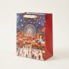 Bolsa de regalo navideña con feria, noria y asas doradas, vista en tres cuartos con lateral rojo visible.