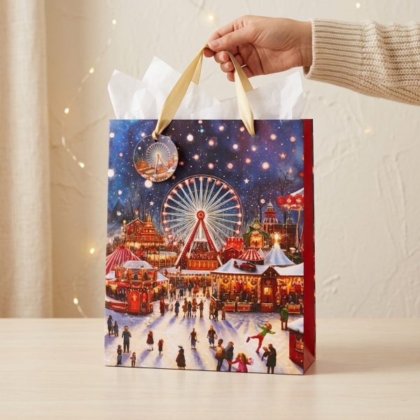 Bolsa de regalo navideña con feria, noria y asas doradas preparada para entregar, con papel de seda decorativo no incluido.